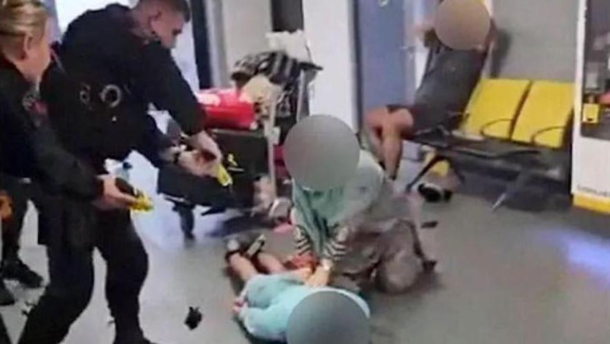 A screenshot of the video where GMP officers after pointing a Taser at a man whose face is blurred out lying face down on the floor during disorder at Manchester Airport. An older woman blurred out has her hands on his back. In the background a man sits with his hands on his head watching.