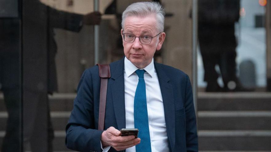 Michael Gove has grey is is wearing glasses, a blue jacket, white shirt and blue tie. He is holding a mobile phone