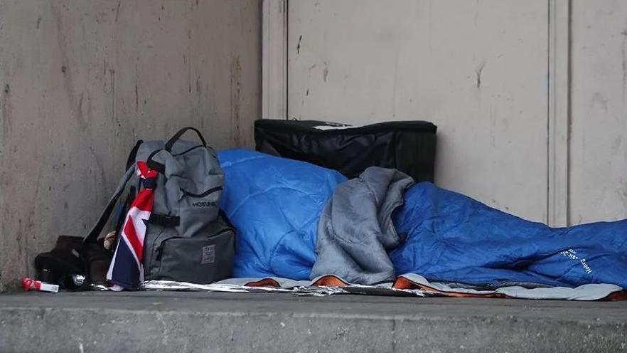 A person inside a blue sleeping bag on a concrete floor. The sleeping bag is on newspaper and there is a grey kitbag with a small Union Jack tied to it leaning against it and an empty cigarette packet on the floor next to the bag.