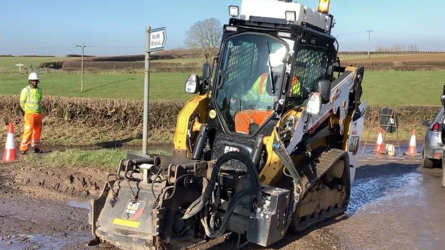 A large yellow machine being driven by a man in flourescent clothing on a muddy road.