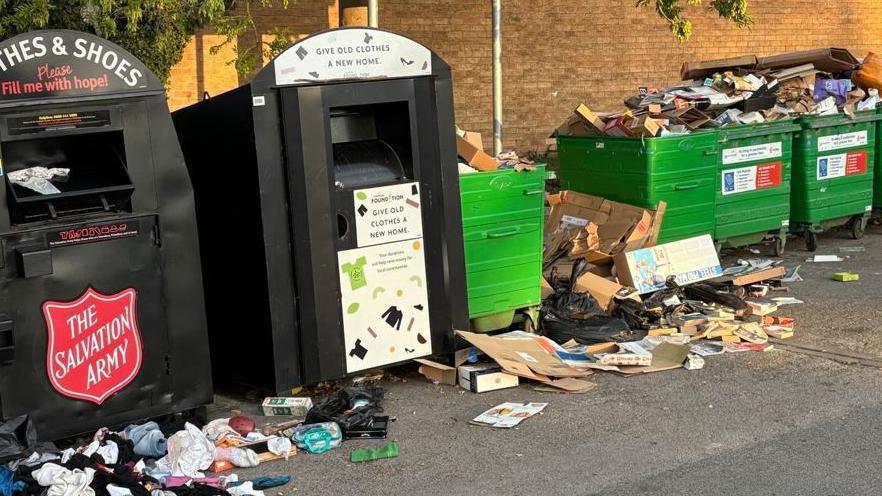 Black and green recycling bins standing against a brick building are overflowing with rubbish. Much of it on the asphalt floor of a car park.