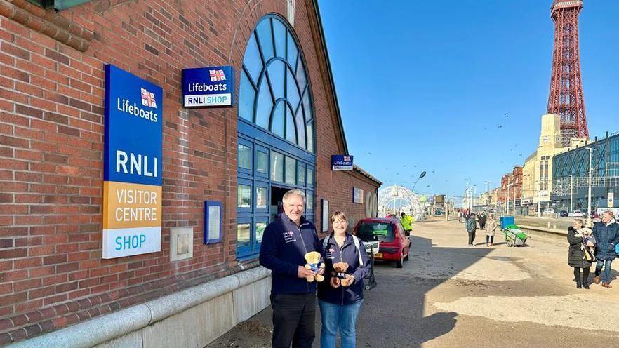 Allan and Helen Thornhill standing outside the RNL lifeboat station in Blackpool with the seafront promenade and Blackpool tower behind them 