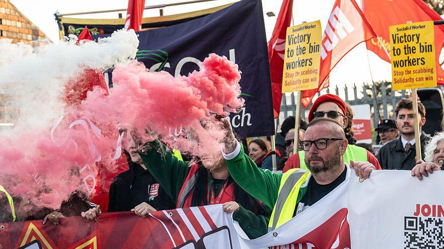 A large protest. A man can be seen holding a banner. He has a bald head and is wearing glasses. He is holding a red flare. There are people all around him holding placards. They look suitably determined.