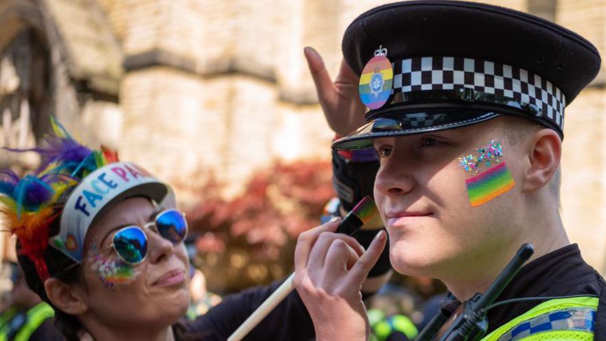 A police officer wearing a black hat with a rainbow badge on it is having his face painted with rainbow stripes during a Pride event.