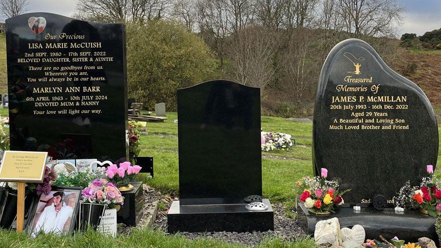 A photo of three graves. The middle one is blank. The left hand grave is Lisa's. It is tall black marble with a small picture of Lisa at the top. There are flowers and photos at the base. The right hand grave is James's. It is black marble with more rounded edges. It also has flowers in bouquets around the plinth.