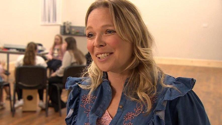 Singer/songwriter Hayley McKay smiles and looks relaxed. She wears a frilled denim blouse. In the background children involved in the project are sitting down playing instruments.