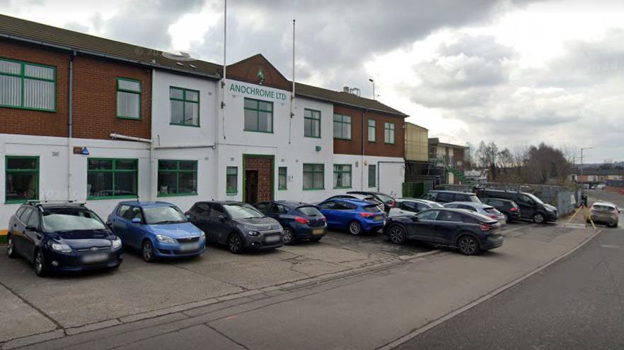 The exterior of Anochrome Ltd, Walsall, which has a brick red and white fascia, with a series of cars parked outside alongside a road running past