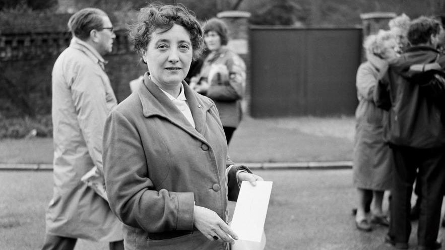 An archive picture of Olive Gibbs from 14 April 1963 at the gates of Windsor Castle. It is during the anti-nuclear weapons march from Aldermaston to London over the Easter weekend. She is about to present the petition. She is looking at the camera and holding a piece of paper. There are people walking behind her.