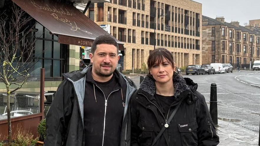A man and a woman stand outside a restaurant on a busy street. It has been raining and the road is wet, both are looking directly into the camera and have dark hair.