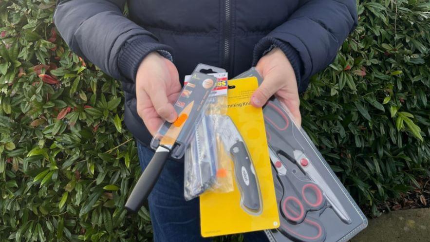 Man wearing dark blue jeans and blue puffer jacket holding several bladed articles including a kitchen knife, Stanley knife and pack of three scissors   