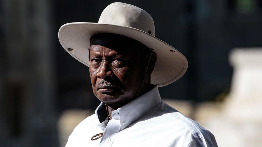 President Yoweri Museveni, wearing a white shirt and a cream wide-brimmed sunhat, looks at the camera unsmilingly