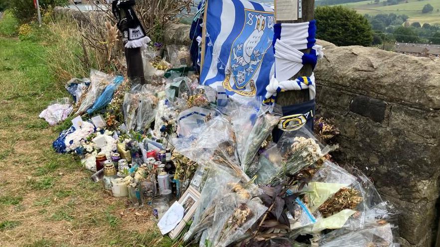 A Sheffield Wednesday flag attached to a wall, with dozens of bunches of flowers in front of it. Drinks cans are also there.