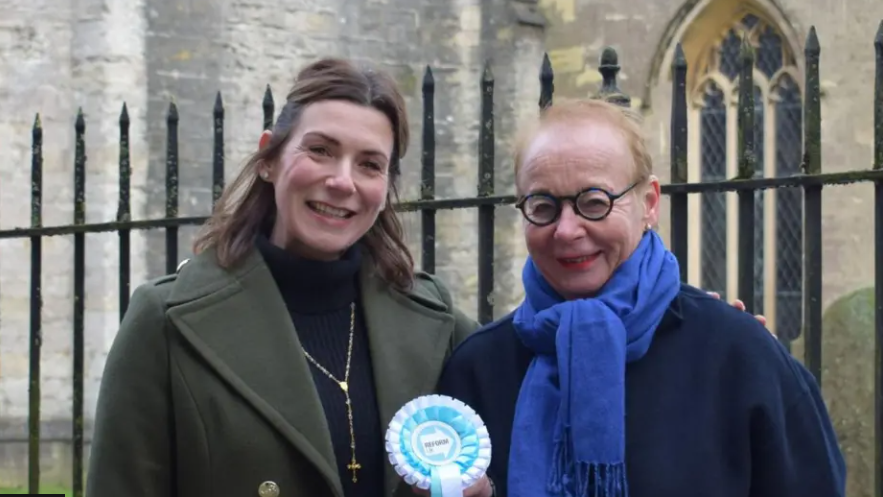 A close-up head and shoulders shot of Aimee and Bente smiling to camera and standing in front of railings with a church in the background. Bente is holding a Reform UK rosette in turquoise and white colours