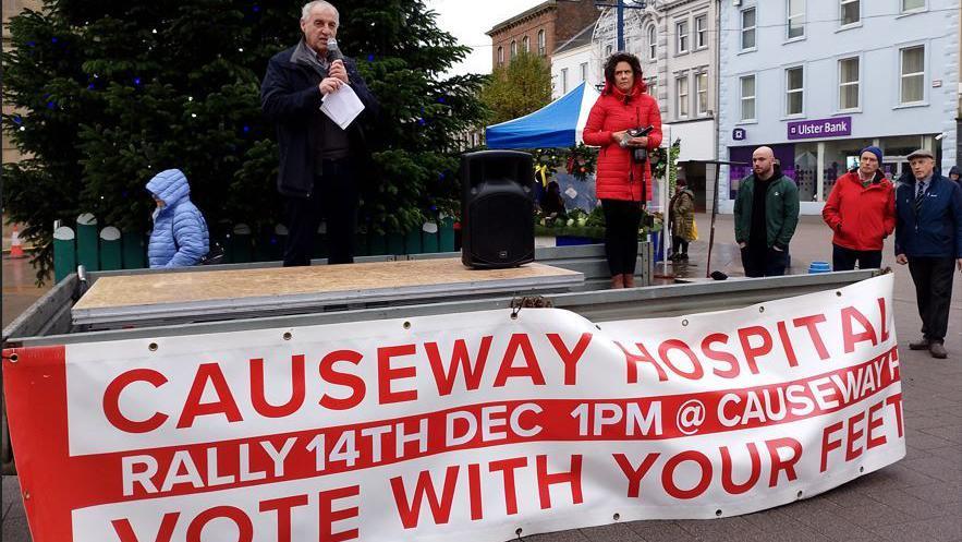 A man and a woman standing on top of a stage with a banner on the front of the stage that reads: Causeway Hospitl, rally 14th December. Vote with your feet".