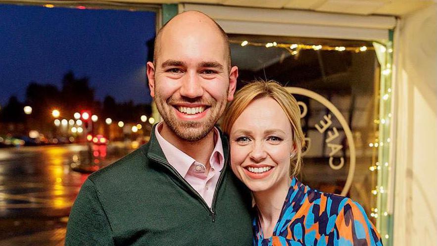 William Seymour dressed in a green jumper in a bar with his wife, wearing orange and blue dress