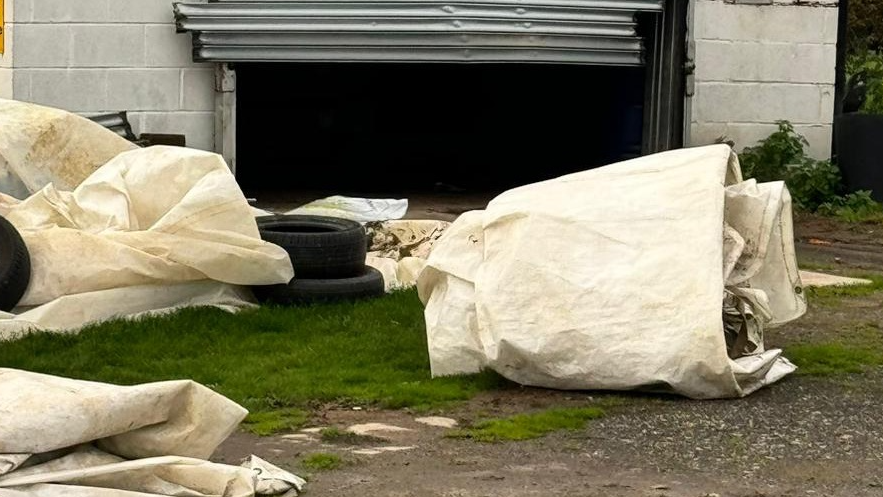 Tyres and sheeting are scattered across grass and gravel in front of an outbuilding with a damaged door.