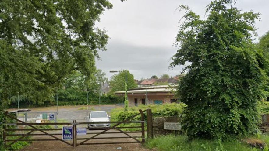 A single storey building set in land. There is a temporary metal fence around the land. A car is parked in front of the fence. A set of gates are closed in front of the car.