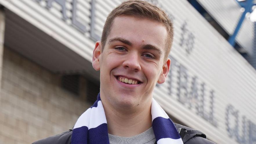 Will Kilgannon in a blue and white striped football scarf. He is standing in front of Millwall Football Club's home ground.