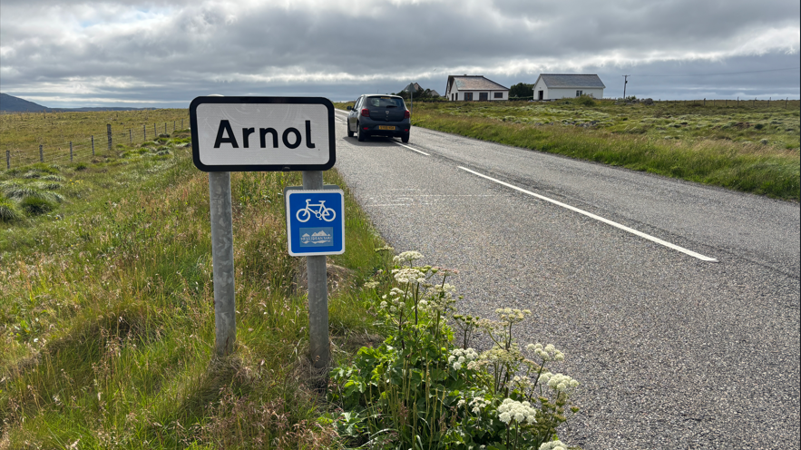 Soidhne baile Àrnoil ri taobh an rathaid. Tha càr a' siubhal air an rathad.