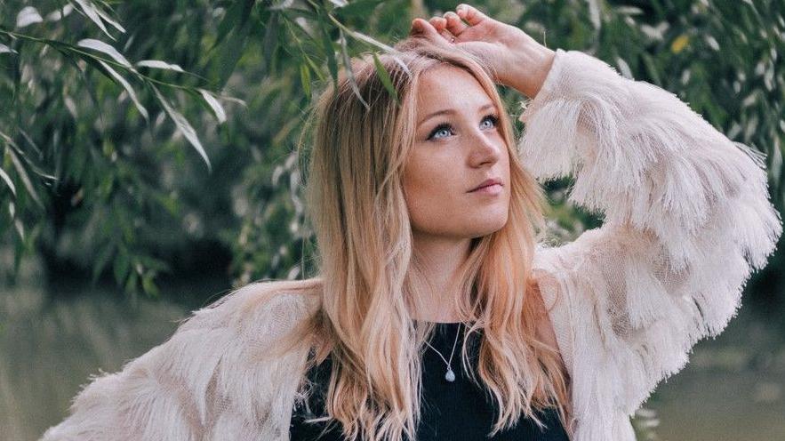 Dolly Mavies next to a tree, the leaves touching her head. She has long fair hair and blue eyes. She wears a black top with a white, feathery jacket.