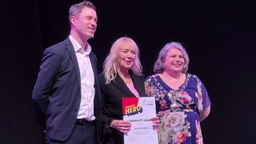 A man and two women stand on a stage. Laura Douglas is in the middle holding her certificate.