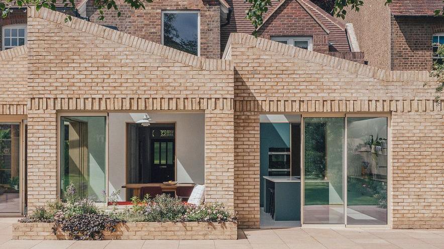 Single-storey brick building in light beige. There are two rooms visible, each with a triangular roof rising to the left. There are tall windows and doors on each and a small flower bed is visible in the foreground in front of the left room.