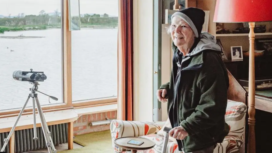 Bettie in outdoor gear and woollen hat, smiling to camera, standing in a room overlooking a lake with a telescope in the window