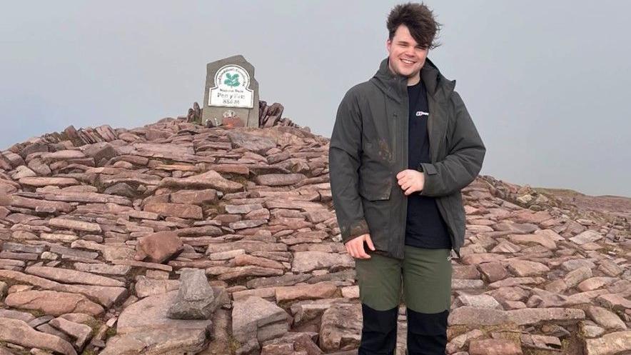 Jordan standing at the summit of Pen Y Fan in a coat