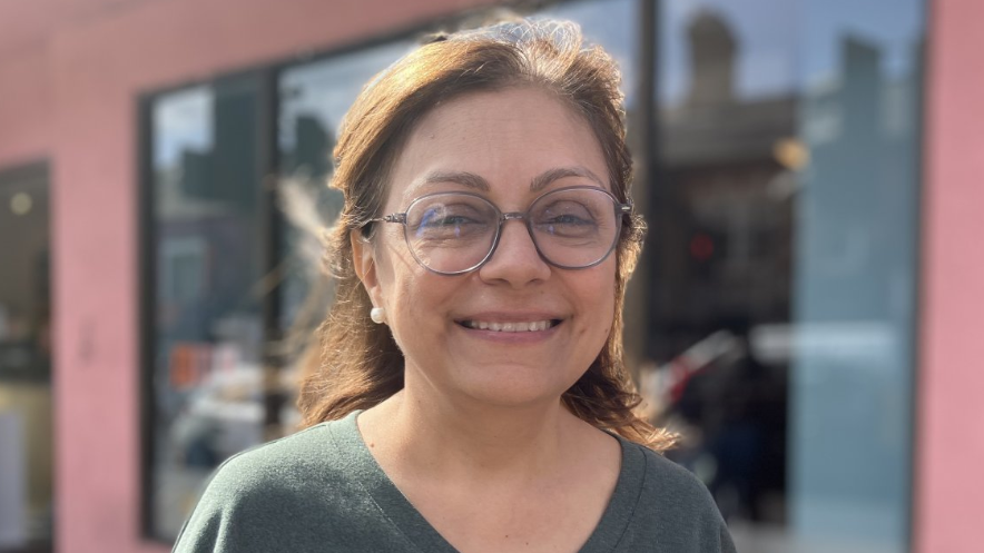 A woman in glasses and a grey top with a deep v neck smiles at the camera. Behind her is a blurred background that looks like a shop window