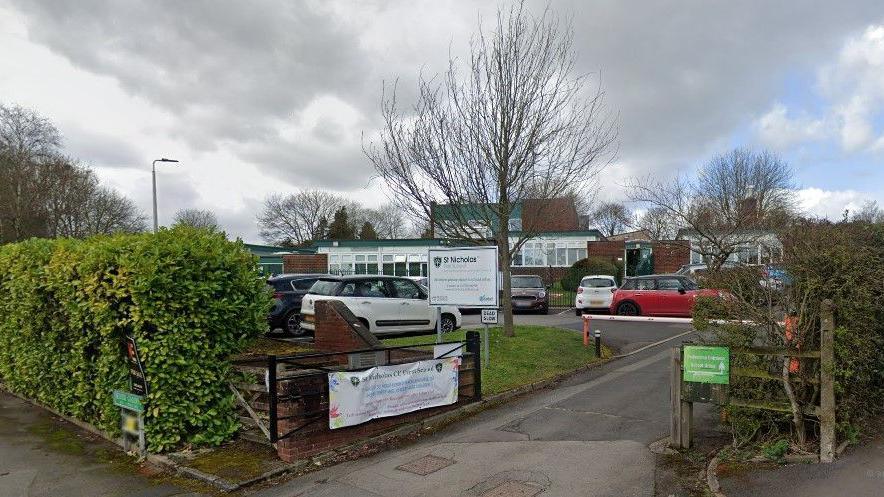 The entrance to a school with a number of cars parked in a small car park bounded by hedges. There is a white sign with black writing that identifies the school and a banner on a gate.