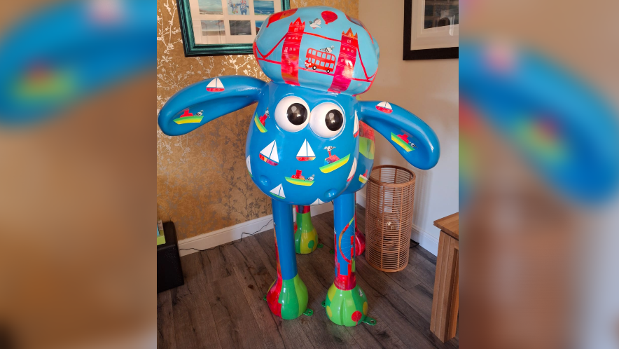 A Shaun the Sheep shaped figure - life-size - in the corner of a living room, painted with lots of blue and motif representing London like Tower Bridge, a red bus, the London Eye and boats.