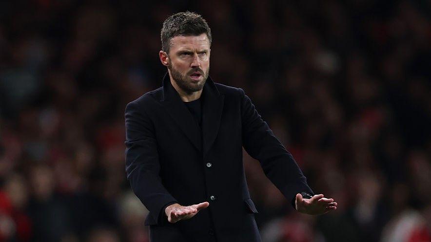 Manchester United interim head coach Michael Carrick makes a palms-down gesture that appears to be calling for calm as he wears a dark coat over a dark top