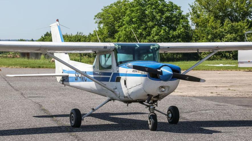 A small plane that is white with blue stripes and has a propeller on the front. It is on a runway in front of trees on a sunny day.