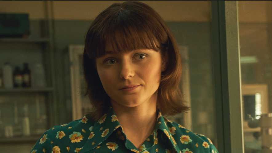 Thomasin McKenzie looking at the camera. She has brown hair and blue shirt with sunflowers on it. She is stood in a laboratory.