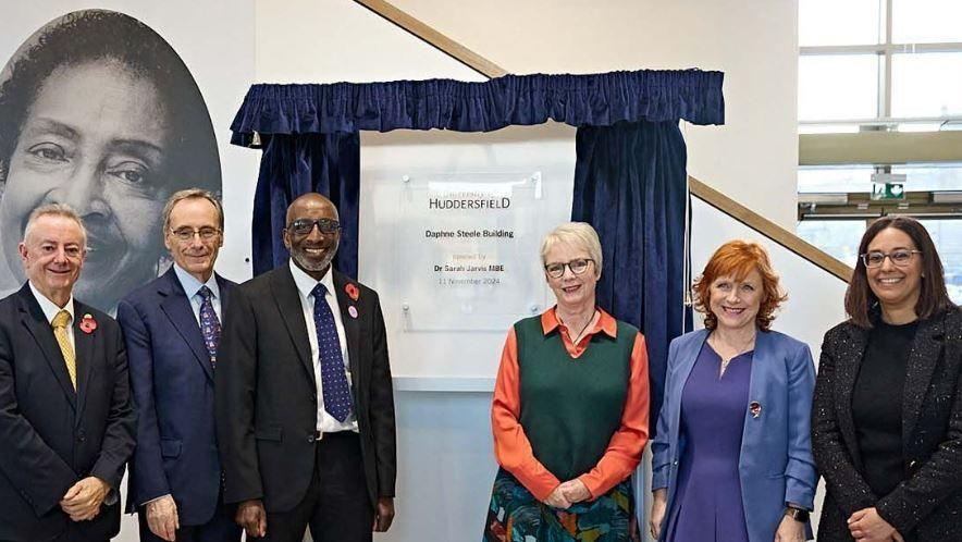 Left to right at the formal opening of the Daphne Steele building:  Professor Bob Cryan (University of Huddersfield Vice-Chancellor), Sir George Buckley (University of Huddersfield Chancellor), Robert Steele, Karin Smyth MP, Dr Sarah Jarvis, Harpreet Uppal MP. To the left is a black and white photo of Daphne Steele, and in the middle is a plaque with the name of the building, between blue curtains.
