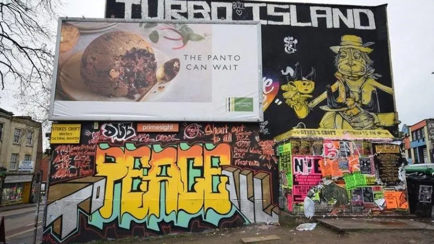 An advertising board shows a Christmas pudding, behind it is lots of graffiti. Some rubbish is left on the land in front.