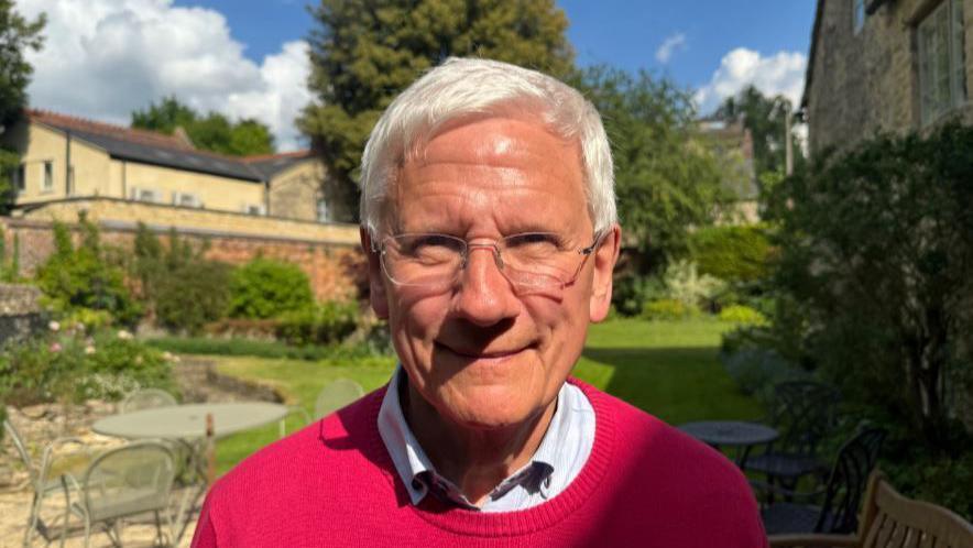 Colin Fletcher smiles for a photo taken outside his house. He has neat white hair and glasses. He is wearing a white collared shirt with a red round-necked pullover on the top. His lawn and garden furniture can be seen in the background.