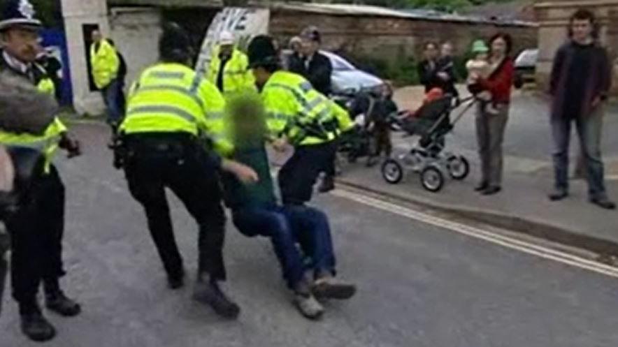 The police evicting squatters from the site in 2006. Two police officers are seen dragging a man. His face is blurred. Another police officer is seen standing aside. Residents are watching them. There are two mothers with prams, holding babies.