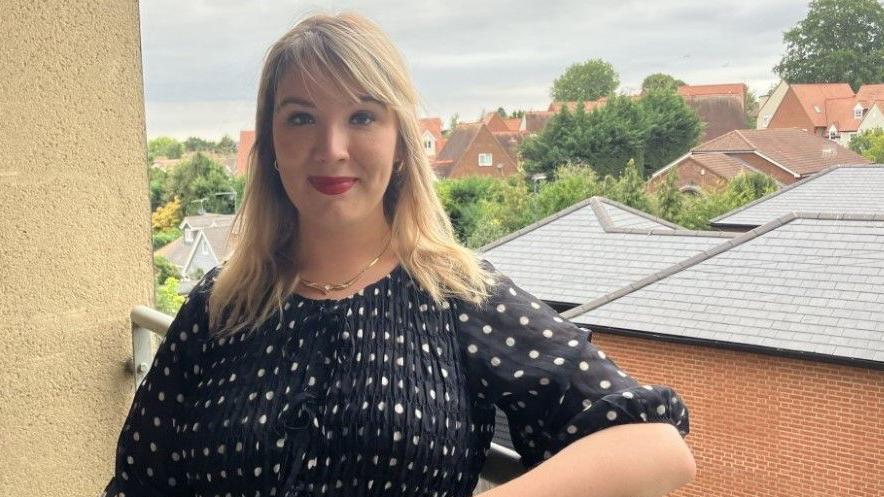 Holly Whitbread is smiling at a camera, standing on a balcony with buildings and rooftops before her. She has long, blond hair and is wearing a black dress with white dots on it.