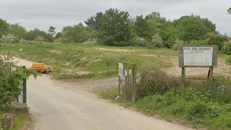 Entry to quarry showing road in past green metal gates, a sign saying "Pury End Quarry" on a board to the right and a green area behind the sign with a track through it - part of the quarried land which has been restored