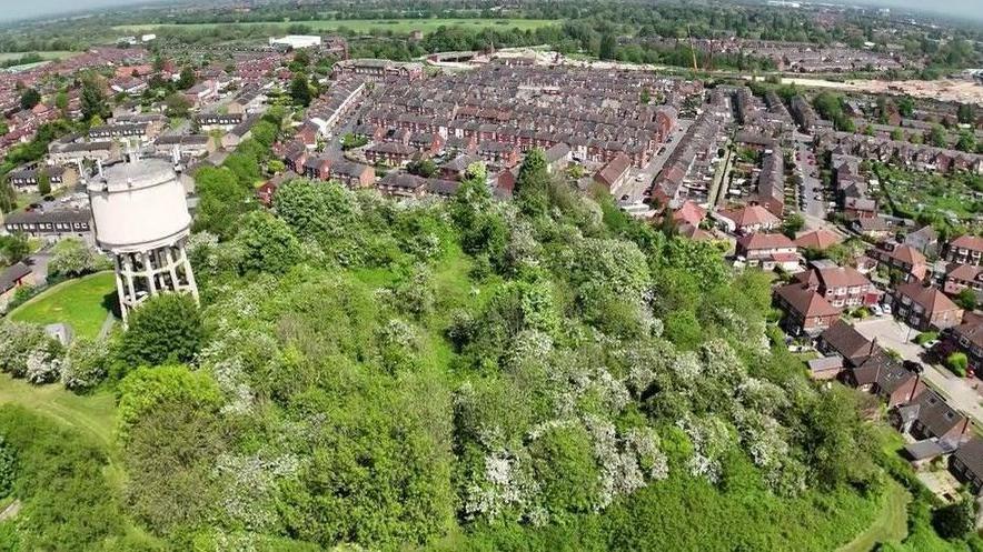 A drone shot showing a mound of grass and greenery with a water tower in the foreground, with streets of houses behind.