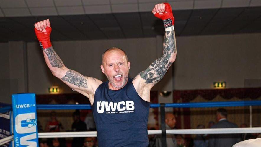 A man stands in a boxing ring with both his arms raised and his mouth wide open as he celebrates. He has red wraps around his hands and wrists, tattoos covering his arms and is wearing a blue tank top. 