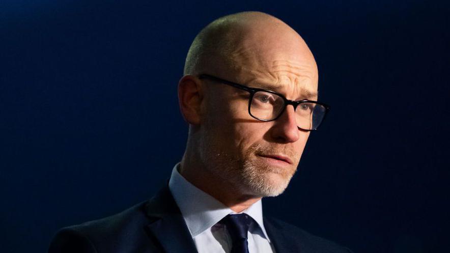 Stephen Kinnock wearing dark glasses and a suit. There is a dark blue background behind him and he has a light shining on his face. He is looking off to the right.