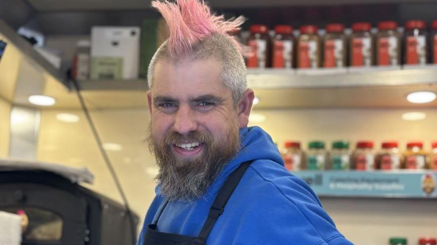 A man with a mohawk and wearing a blue hoodie smiles into the camera. Rows of food jars can be seen on the shelves behind him.