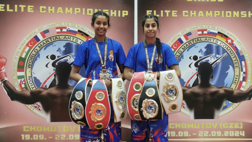 Dheep, 17 and Simran, 14 holding three belts each while standing in front of a kickboxing championship sign