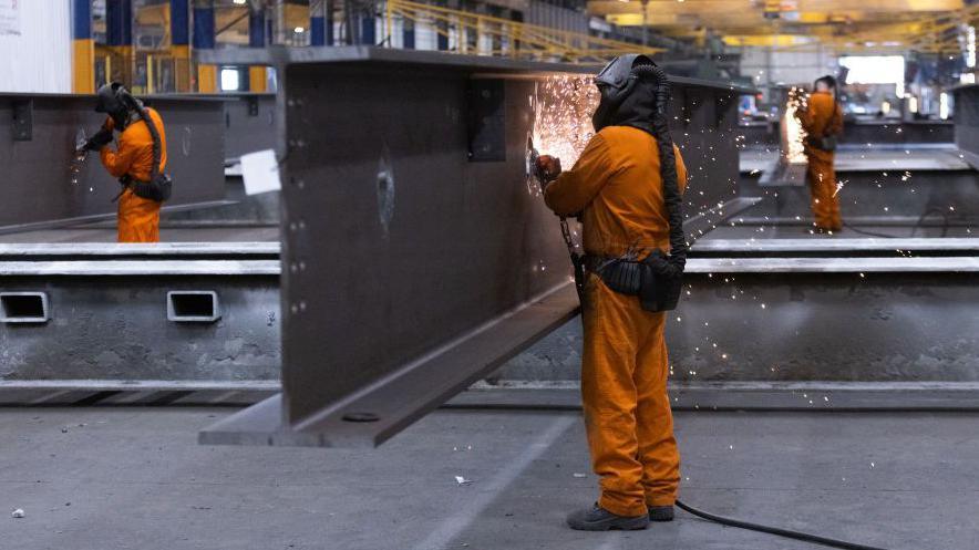 A worker dressed in orange overalls with a protective mask covering his face grinds smooth a section of fabricated steel beam at Severfield Plc steel fabricators in Dalton, near Thirsk. There are two workers in similar clothing in the background of the image.