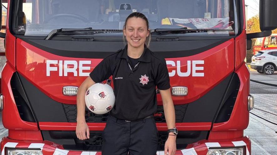 A woman, Bow Jackson, wearing black firefighters' uniform, with a football under her right arm, standing in front of a red fire engine