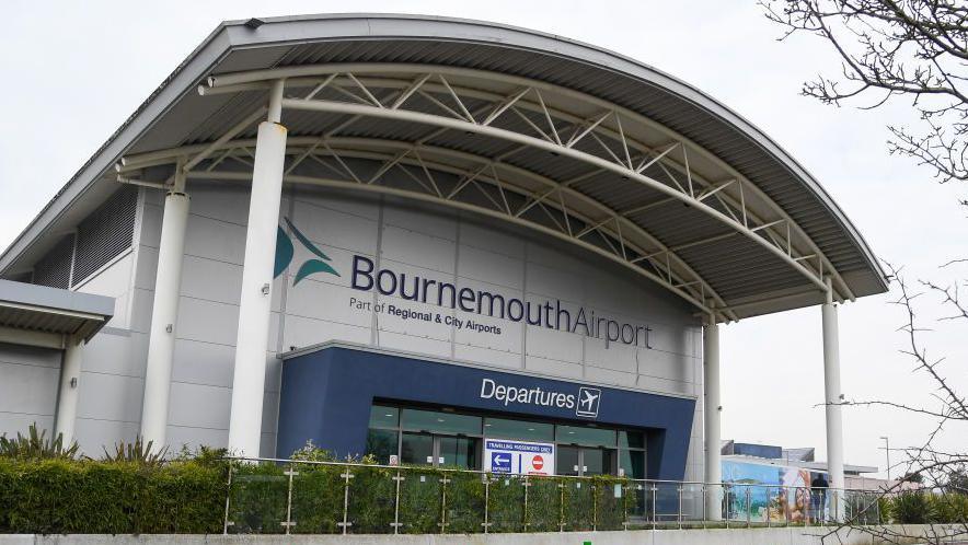 Bournemouth Airport Departure building - a grey building with a large portico held up by a number of columns and a blue surrounding to the glass entrance and a green hedge at the front.