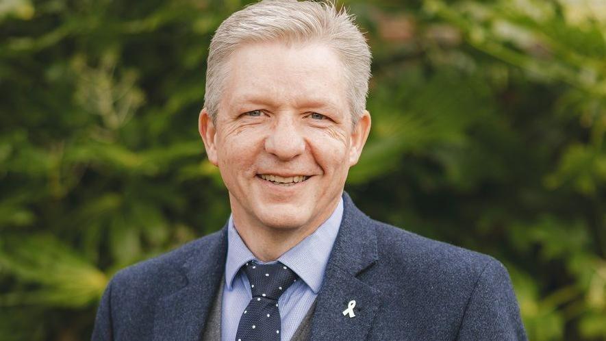 A man is stood looking at the camera wearing a blue jacket and blue tie. He has greenery behind him and is smiling. He is smiling.