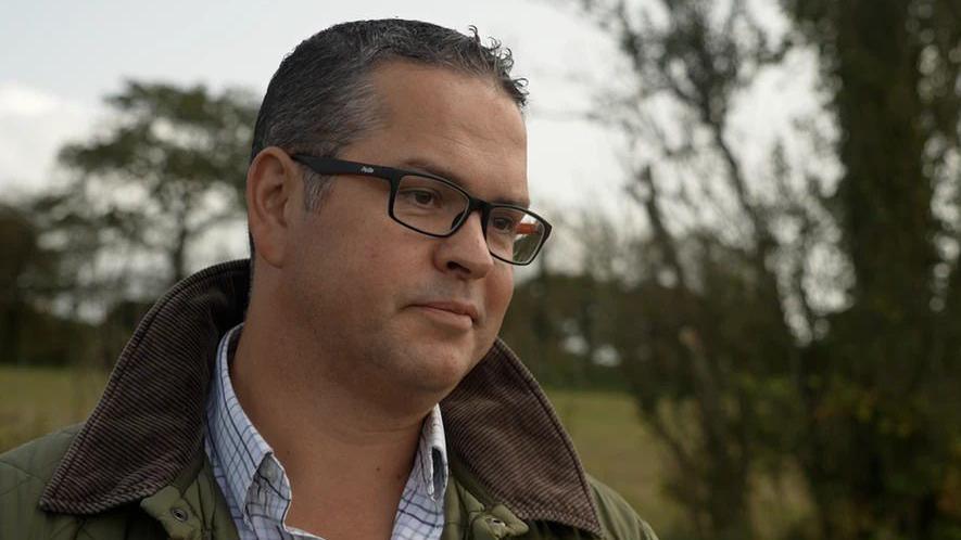 Marcus Calvani is a white man with short, dark hair. He is wearing a checked shirt and a quilted green jacket with a corduroy collar. He is wearing black square-frame glasses. He's in the left of the frame looking towards the right. The background is out of focus trees and grass.